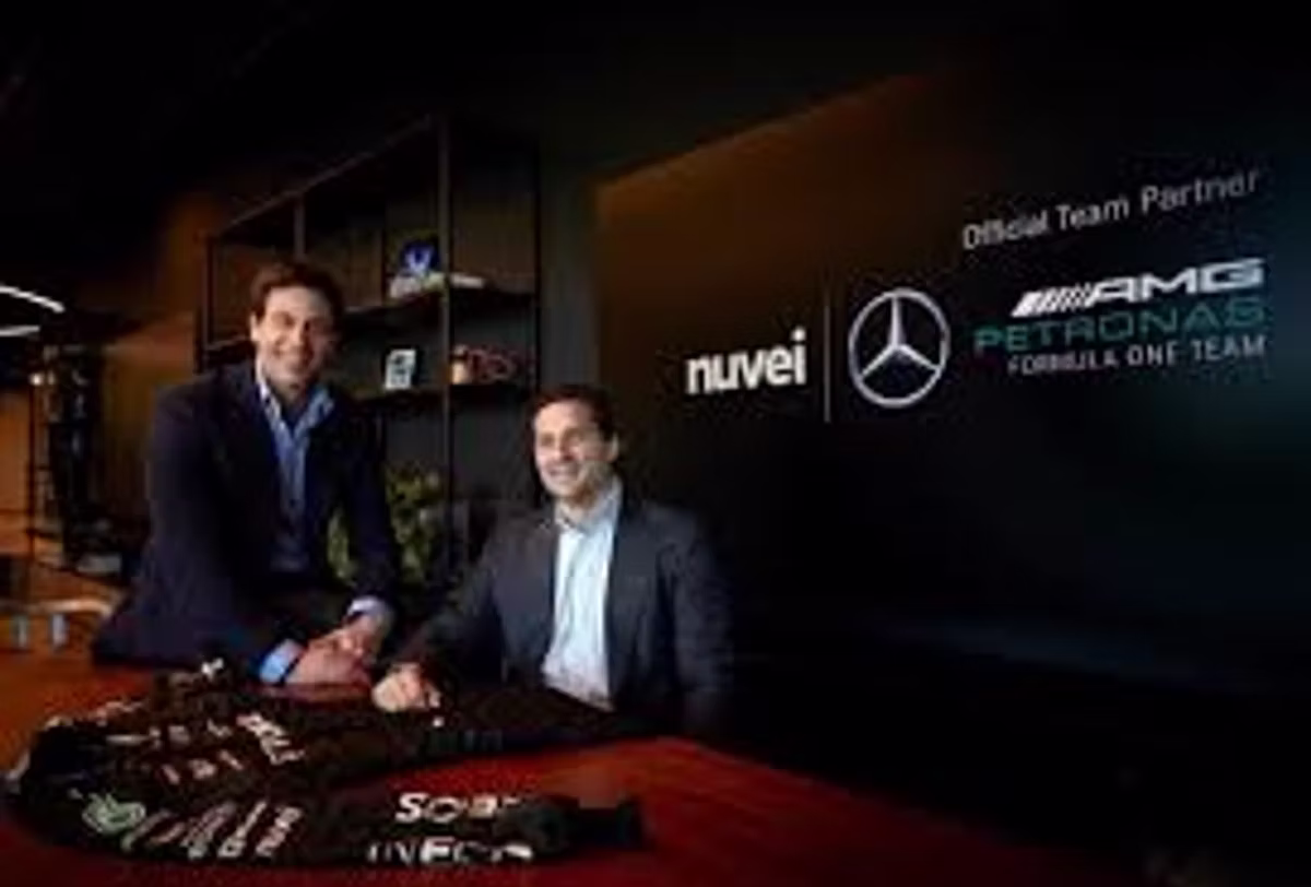 Two men seated at a table with a signed team jacket in front of them, beside a screen reading "Official Team Partner" with the Nuvei and Mercedes-AMG Petronas Formula One Team logos.