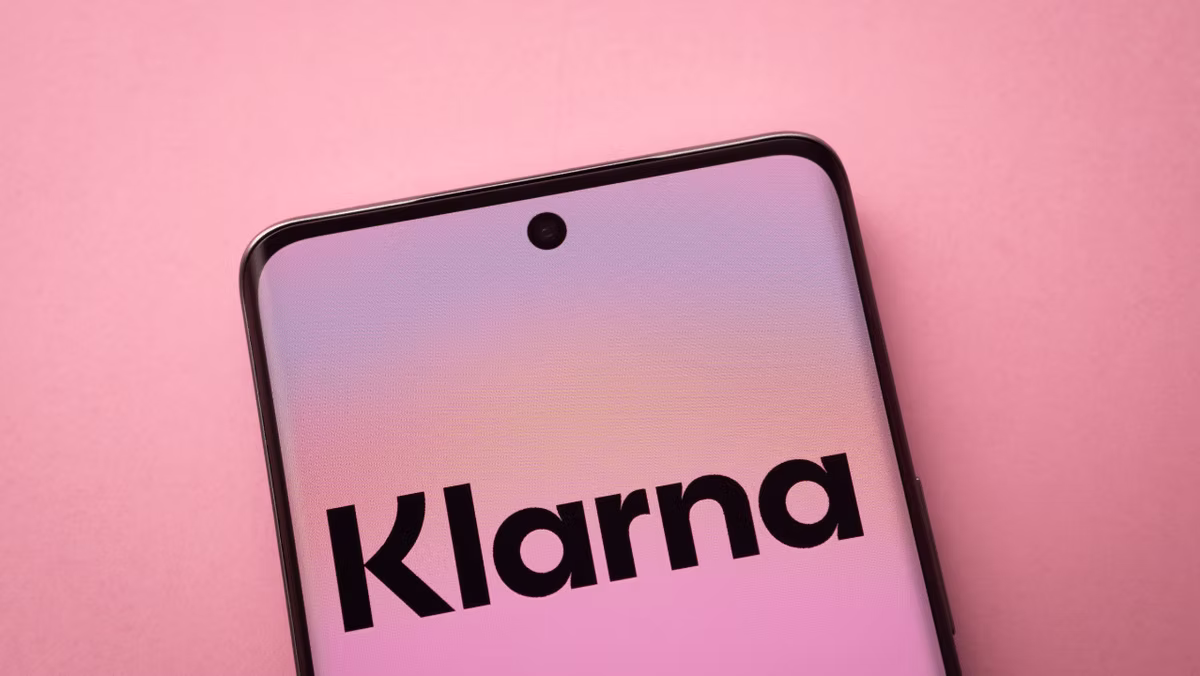 Smartphone in close-up on pink background, screen displays "Klarna." Soft pastel colors evoke a calm, modern feel.